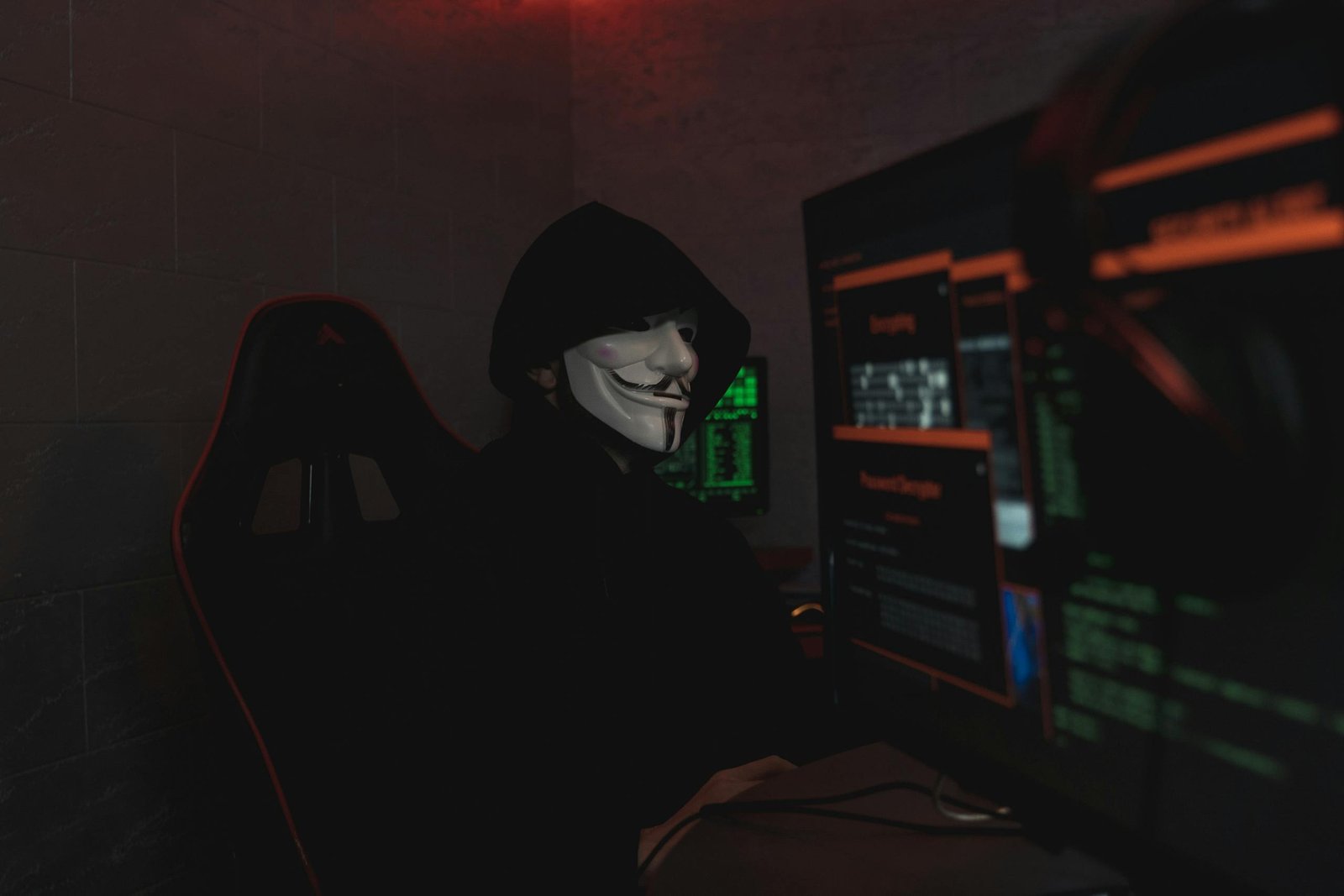 An anonymous hacker wearing a mask working on a computer in a dark room.